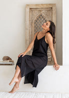 Woman sits in black cotton voile maxi slip with V-neckline, adjustable spaghetti straps and side splits