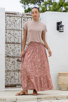Woman wearing a pink top and rose pink based floral skirt standing outdoors against a white wall.