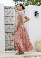 Woman wearing a pink top and floral skirt standing in front of a white wall with a decorative pattern.