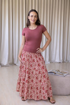 Woman wearing a pink top and floral skirt standing indoors with a curtain background