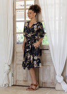 Woman wearing over the knee length wrap dress in black based cotton print and a hand in pocket.