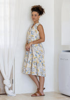Woman stands sideways wearing a sleeveless neutral floral cotton dress with pockets, mandarin collar and a two tiered skirt.