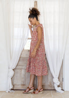 Woman stands sideways wearing a sleeveless rose pink floral cotton dress with pockets, mandarin collar and a two tiered skirt.