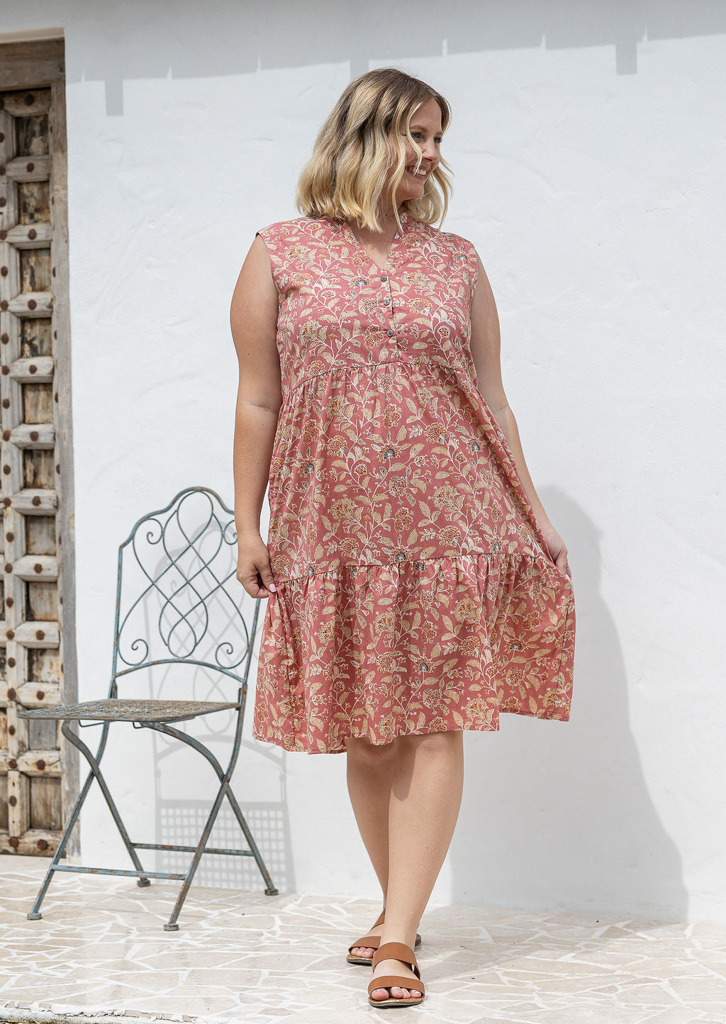 Woman wearing a rose pink based floral sleeveless, knee length dress with a buttoned bodice, tiered skirt and pockets. 