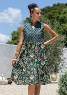 Woman wearing a knee length cotton sleeveless dress with buttoned bodice and  two tiered skirt in green based floral print.