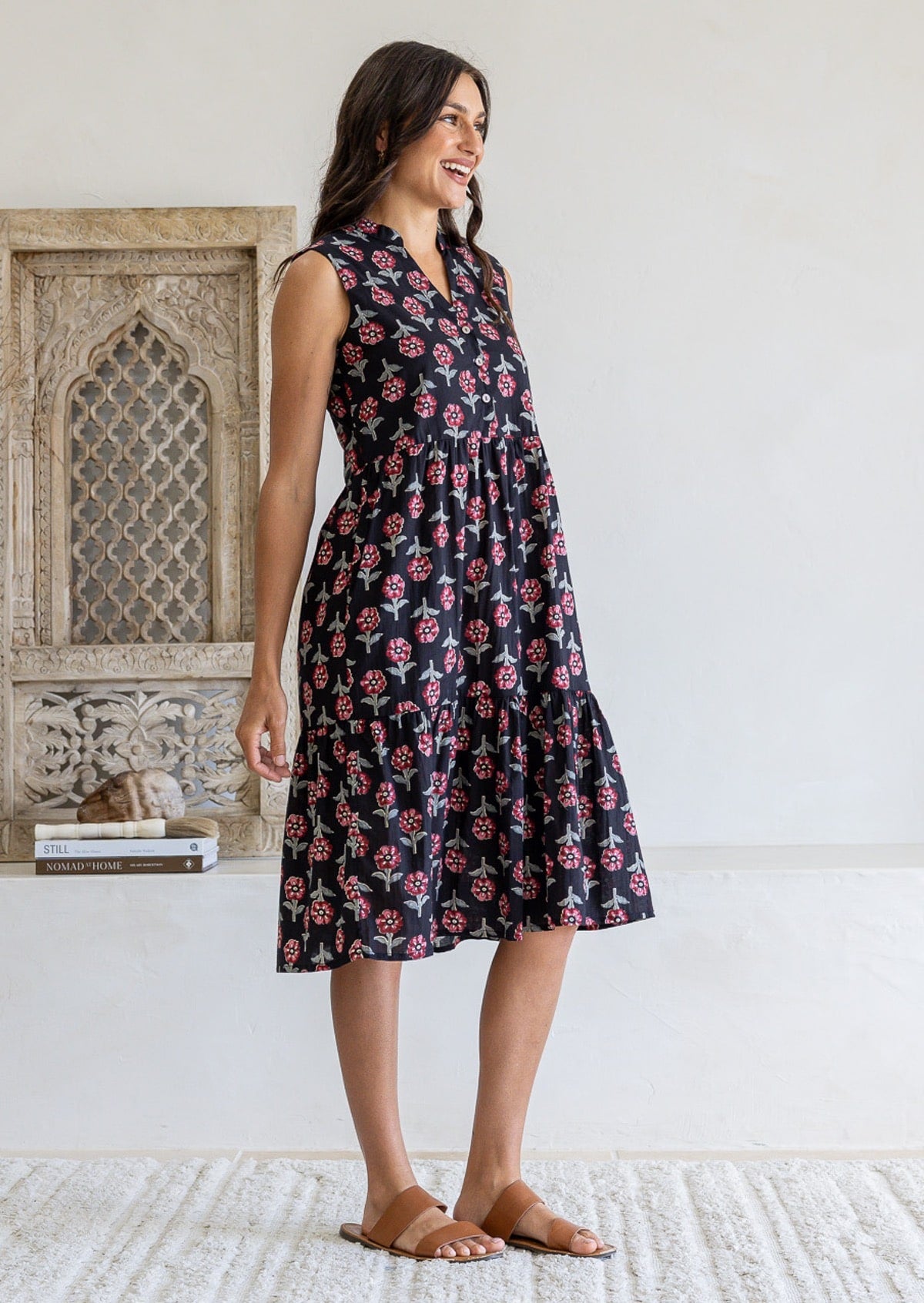 Woman stands sideways wearing a sleeveless black floral cotton dress with pockets, mandarin collar and a two tiered skirt.
