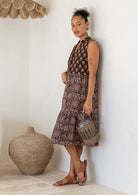 Woman in a brown 100% cotton patterned dress holding a woven basket against a white wall.