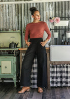 Woman standing in a rustic kitchen wearing black cotton corduroy wide leg pants and a dusty pink top, with hands in pockets