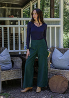 Woman standing on a porch wearing a dark blue top and green pants.