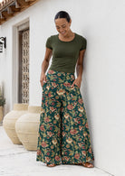 Woman wearing a green top and wide legged green cotton floral pants standing against a white wall.