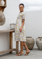 Woman stands sideways wearing a 100% cotton dress in neutral based floral print with short sleeves