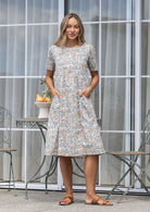Woman poses in a white based cotton floral short sleeve dress with round neckline and hand in pockets