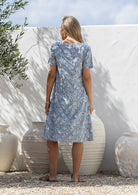 Woman stands backwards wearing a 100% cotton dress in blue based floral print with short sleeves. 