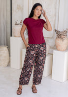 Woman standing in front of white wall wears 100% cotton wide leg Indian traditional print pants in black with elastic waist, pockets and pleated ankles