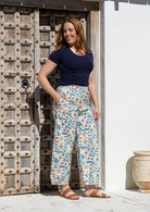 Woman standing wears 100% cotton wide leg floral print pants in white with elastic waist, pockets and pleated ankles with navy blue t-shirt top in front of old wooden door