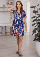 Woman poses in a 100% cotton bold botanic printed dress V-neckline short sleeve gathered waistline above the knee skirt.