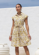 Woman wearing a 100% cotton yellow floral print shirt dress with collar, cap sleeves, and tie waist. 