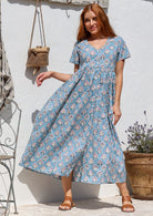 Woman standing in a 100% cotton floral print dress with a V-neckline, short sleeves, pockets, and buttons through to the knee.