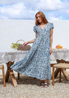 Woman wearing a 100% cotton white based blue floral print maxi dress with V-neck and cap sleeves. 