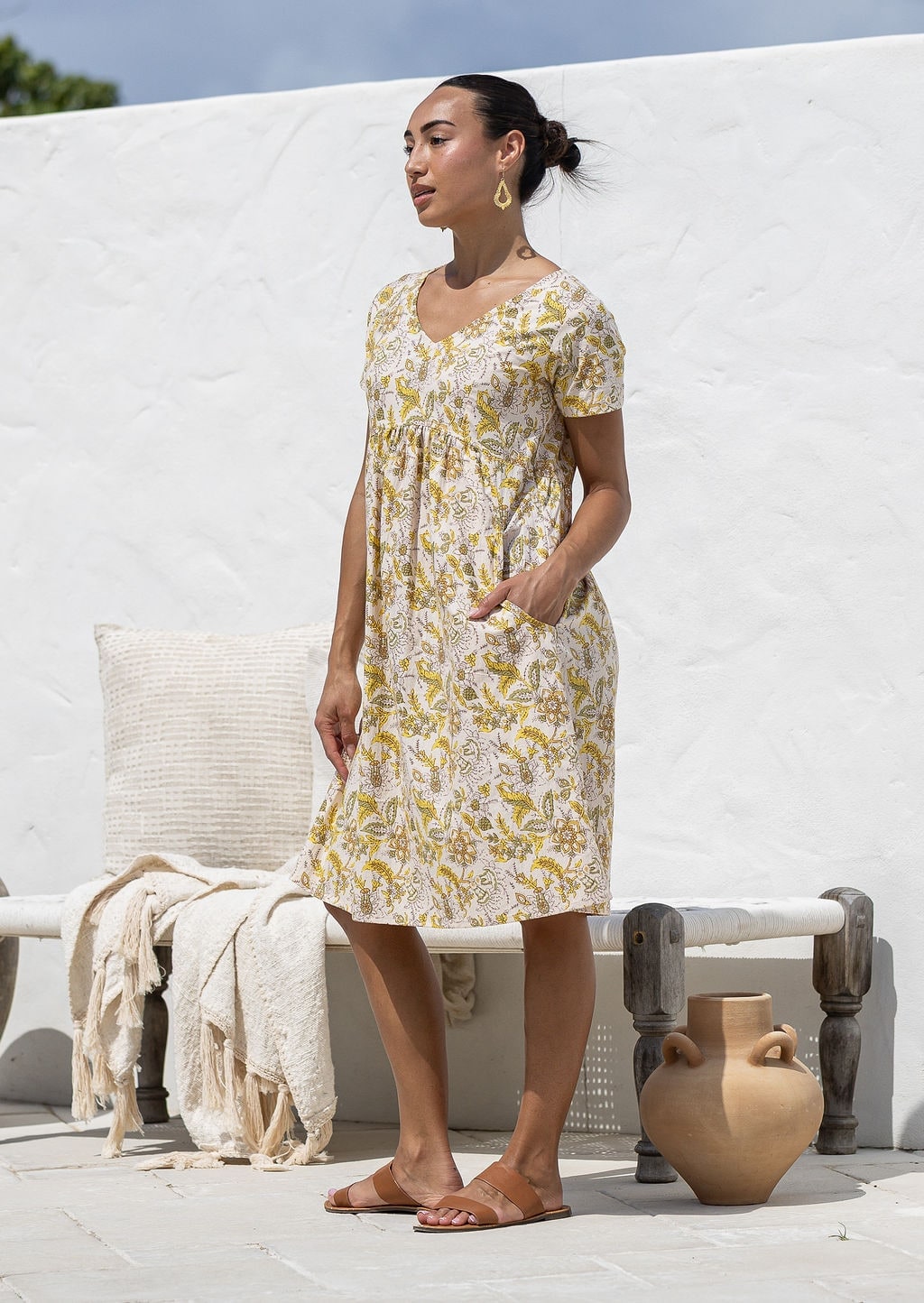 Model stands sideways wearing a cotton V-neck relaxed fit dress with empire waistline and short sleeves with one hand in pocket