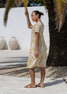 Woman stands sideways wearing a 100% cotton dress in cream based floral print with short sleeves. 