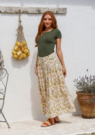 Woman stands sideways wearing a basic top and floral print, shin length cotton skirt with button through front and box pleats.