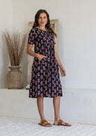 Woman stands sideways wearing a 100% cotton dress in black based floral print with short sleeves and one hand in pocket