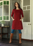 Woman wearing red corduroy A-line dress with 3/4 sleeves with hands in pockets over leggings and boots