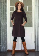 Woman wearing brown corduroy A-line dress with 3/4 sleeves with hands in pockets over leggings and boots