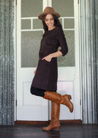 Woman wearing a brown dress, hat, and boots standing in front of a white door with glass panels.