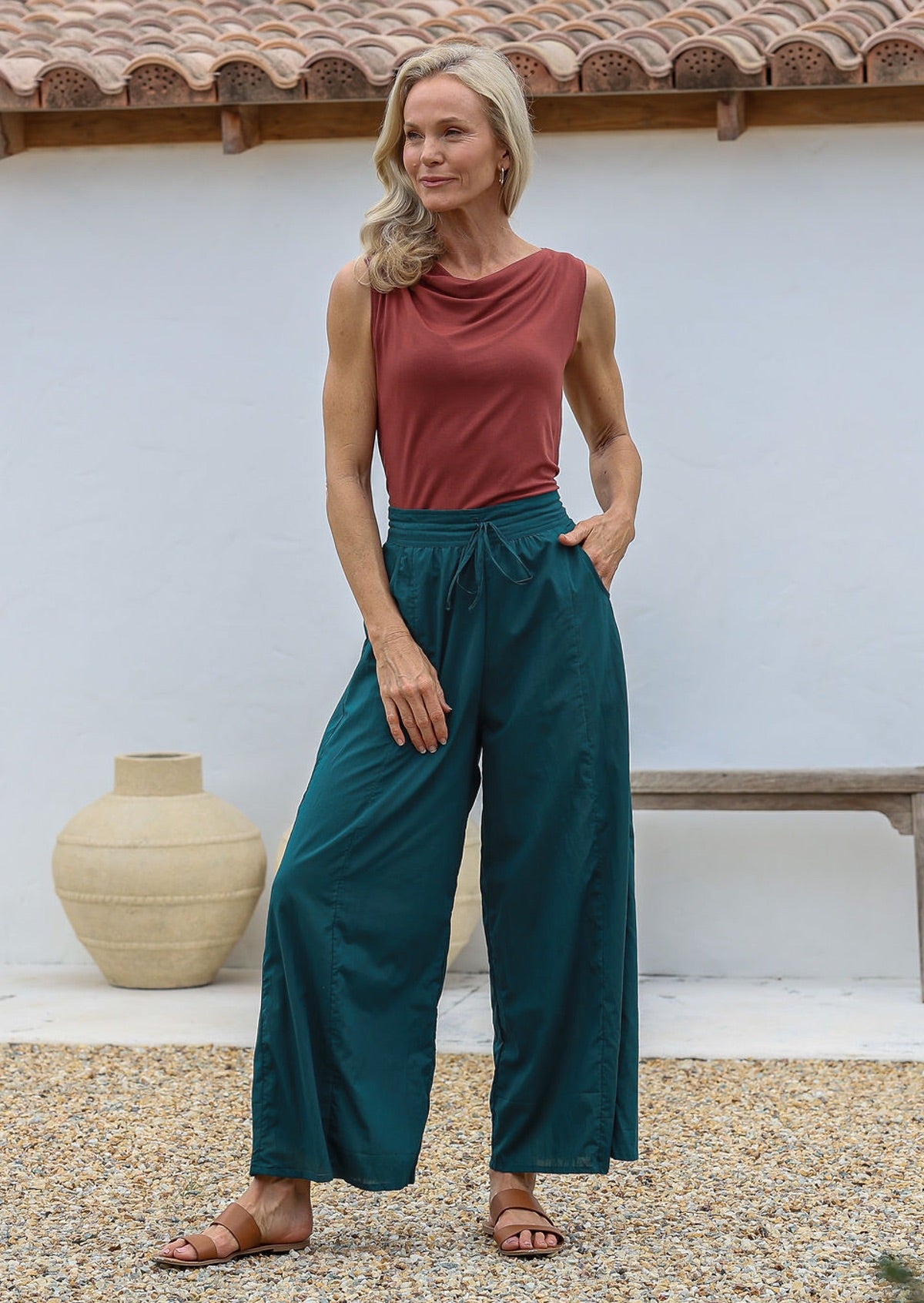 Woman poses in relaxed fit wide-leg cotton pants in deep teal with pockets, elastic waist and drawstring tie.