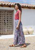 Woman wearing a pink top and rayon pants standing outdoors with a white wall and wooden door in the background.