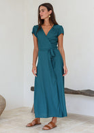 Woman wearing a teal rayon wrap maxi dress standing in a minimal indoor setting.