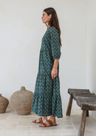 Woman standing in front of wooden bench wears three tiered cotton maxi dress with 3/4 sleeves, pockets and mandarin collar