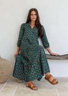 Woman sits on a wooden bench posing in lightweight cotton floral printed maxi dress with 3/4 sleeves, mandarin collar and buttoned bodice