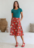Woman wearing a basic green top and red floral a-line cotton skirt with side zipper and one hand in pocket.