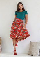 woman leans on wall wearing a knee length floral cotton skirt with a-line shape, pockets and side zipper.