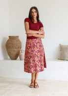 Woman wearing a maroon top and floral cotton skirt standing in a room with a woven basket and pillows.