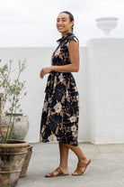 Model stands sideways near wearing a mid length, shirt style cotton dress with black based floral print