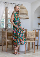 Model stands wearing a mid length, shirt style floral print cotton dress in olive green based floral print with one hand in pocket