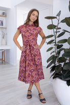 Model stands wearing a midi length floral printed shirt style cotton dress with cap sleeves, buttoned bodice and a removable waist tie