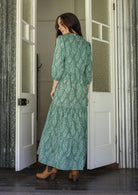 Woman stands backwards wearing a 100% cotton tiered maxi dress in cream based floral print with 3/4 sleeves, pockets and mandarin collar