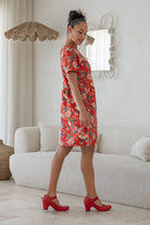 Woman stands sideways wearing a relaxed fit, red based floral cotton print dress with pockets and empire waistline, standing in a living room.