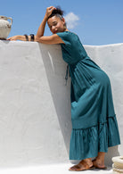 Woman in a teal maxi dress leaning against a white wall with a blue sky.