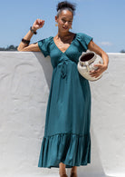 Woman in a teal maxi,  Christmas dress holding a ceramic pot against a white wall with blue sky.