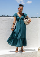 Woman in a teal maxi, perfect Christmas dress holding a ceramic pot against a white wall with blue sky.