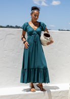 Woman in a teal dress maxi dress holding a ceramic pot against a white wall with blue sky.