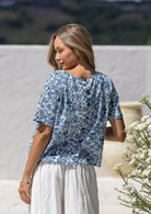 Woman standing backwards wearing a relaxed n fit white based blue floral printed cotton top with decorative buttons down the centre back