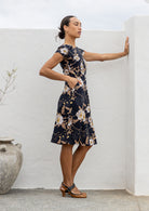 Model with one hand in pocket wears a knee length A line dress with cap sleeves in black based floral print
