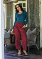 Woman in teal top and red corduroy pants standing on a wooden deck with wicker chairs and cushions.